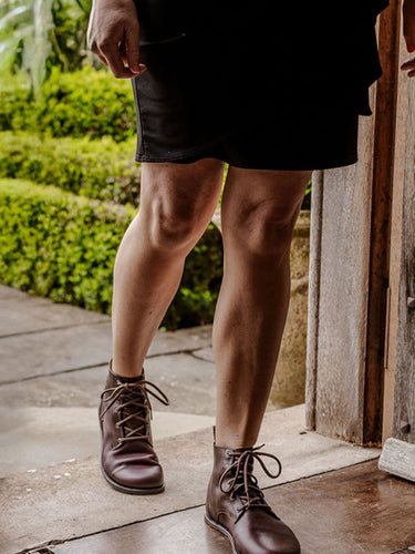 Person wearing a black skirt and goodyear welted barefoot boots in brown leather, standing on a stone path with greenery in the background, best women barefoot dress boots